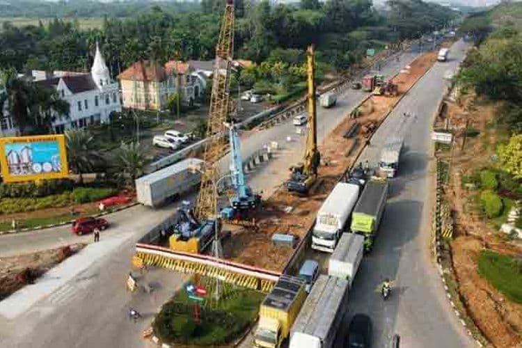 Foto udara lokasi proyek konstruksi jalan layang di median jalan raya urban padat Indonesia, memperlihatkan mobilisasi alat berat crawler crane dan drill rig di area kerja terbatas.